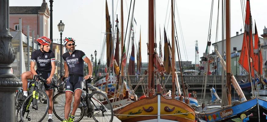 Zwei Radfahrer in Schwarz stehen am Hafen mit bunten Segelbooten