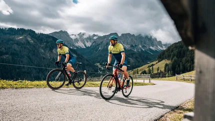 Two cyclists riding uphill on a winding road with mountains in the background