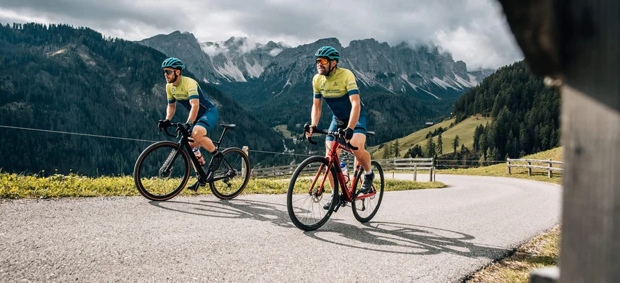 Herbst auf Schotter im Excelsior Dolomites Life Resort****s © Jens Scheibe Zwei Radfahrer fahren bergauf auf einer kurvigen Straße mit Bergen im Hintergrund