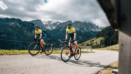 Zwei Radfahrer fahren bergauf auf einer kurvigen Straße mit Bergen im Hintergrund