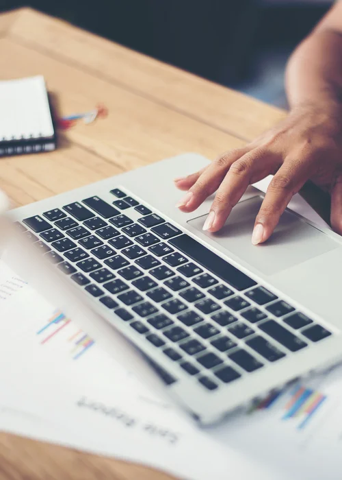 Hand bedient das Trackpad eines Laptops auf einem Holztisch mit Dokumenten