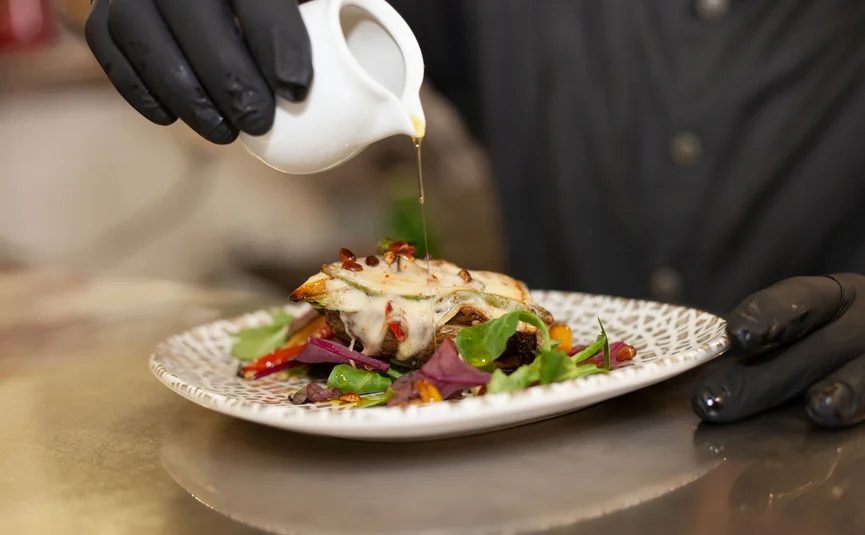 Person mit schwarzen Handschuhen gießt Sauce über ein Gericht mit Gemüse und Käse
