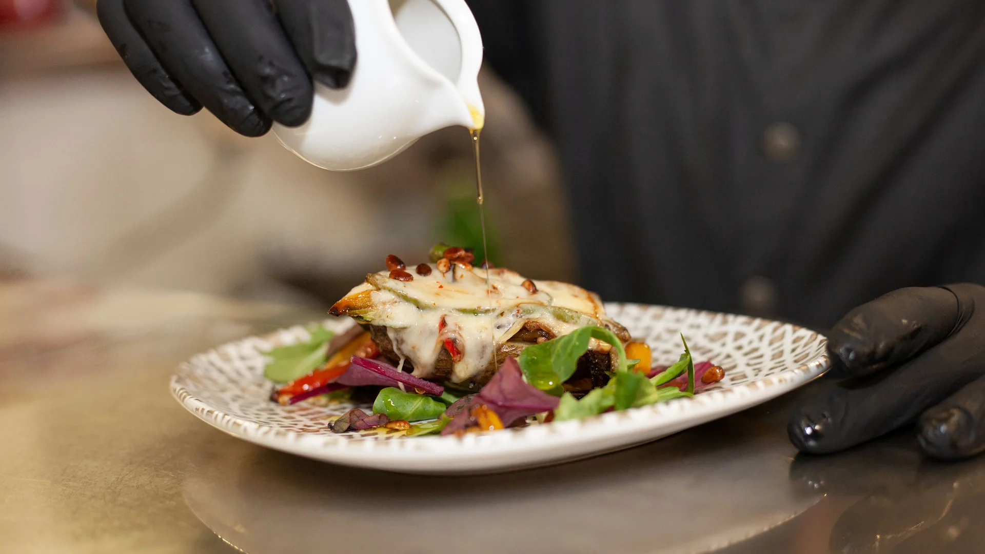 Person mit schwarzen Handschuhen gießt Sauce über ein Gericht mit Gemüse und Käse