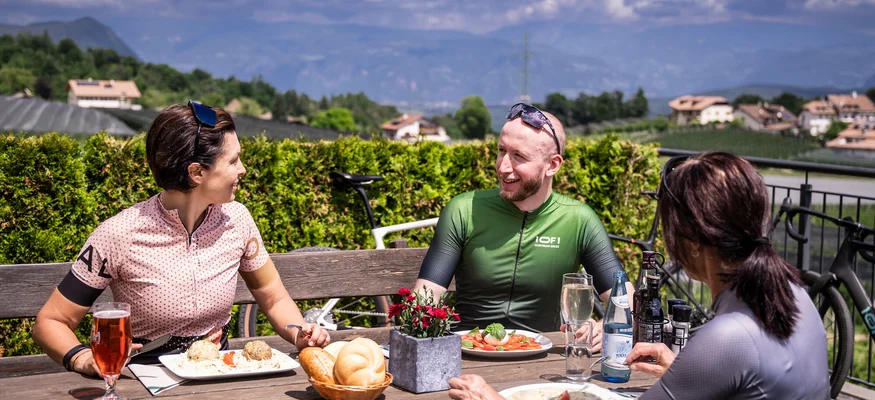 Gravel & Törggelen: Warum Tramin im Herbst perfekt für Gravelbiker ist © Heiko Mandl Drei Radfahrer genießen Essen und Getränke im Freien mit Bergblick