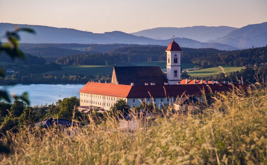 Stift St. Georgen **** © Elias Jerusalem Klostergebäude mit rotem Dach vor Berg- und Seenlandschaft bei Sonnenuntergang