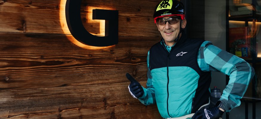 Gravelbiken im Savinja-Tal © Ziga Intihar Mann mit Fahrradhelm zeigt Daumen hoch vor beleuchtetem G an Holzwand