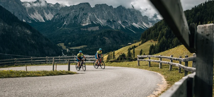Zwei Radfahrer fahren auf bergiger Straße mit Holzabsperrung und bewölkten Bergen