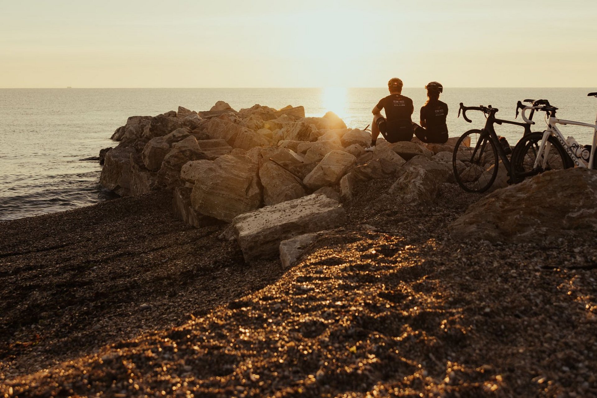 Gravelbike Holidays © Alex Moling Zwei Radfahrer sitzen auf Felsen am Meer und schauen bei Sonnenuntergang aufs Wasser