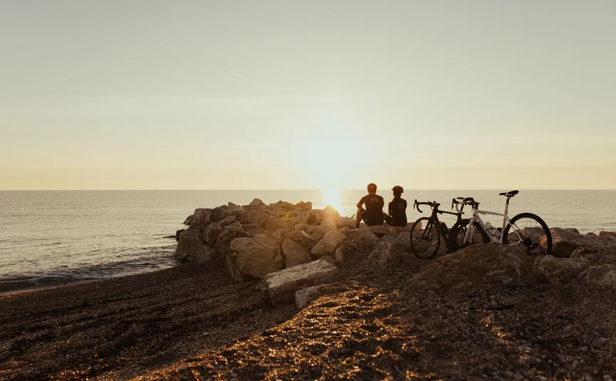 Two cyclists sitting on rocks by the sea watching the sunset over the water