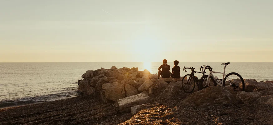 Zwei Radfahrer sitzen auf Felsen am Meer und schauen bei Sonnenuntergang aufs Wasser