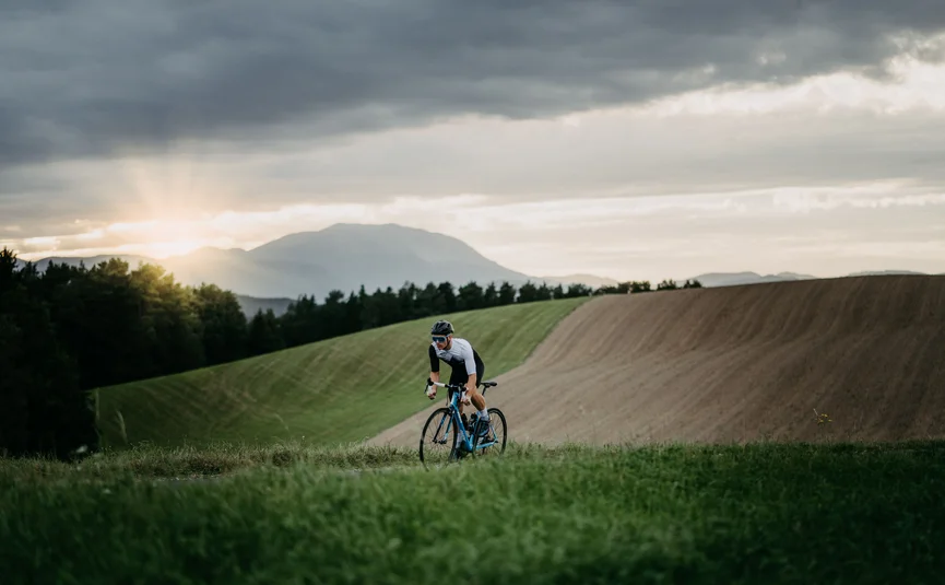 Radfahrer fährt auf Landstraße bei Sonnenuntergang vor hügeliger Landschaft