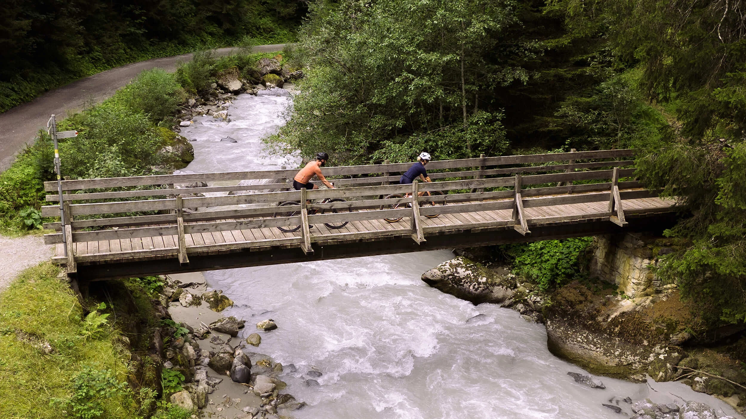 Austria © Janine Brugger - Vorarlberg Tourismus Two cyclists crossing a wooden bridge over a river in a forest