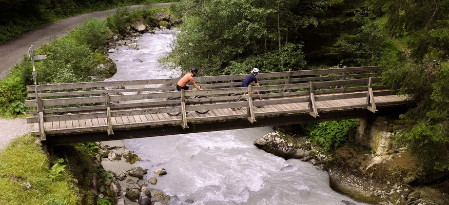 Gravelbiken in Vorarlberg: Die 7 schönsten Highlights zwischen Bodensee und Alpen © Janine Brugger - Vorarlberg Tourismus Zwei Radfahrer fahren über eine Holzbrücke über einen Fluss in einem Wald