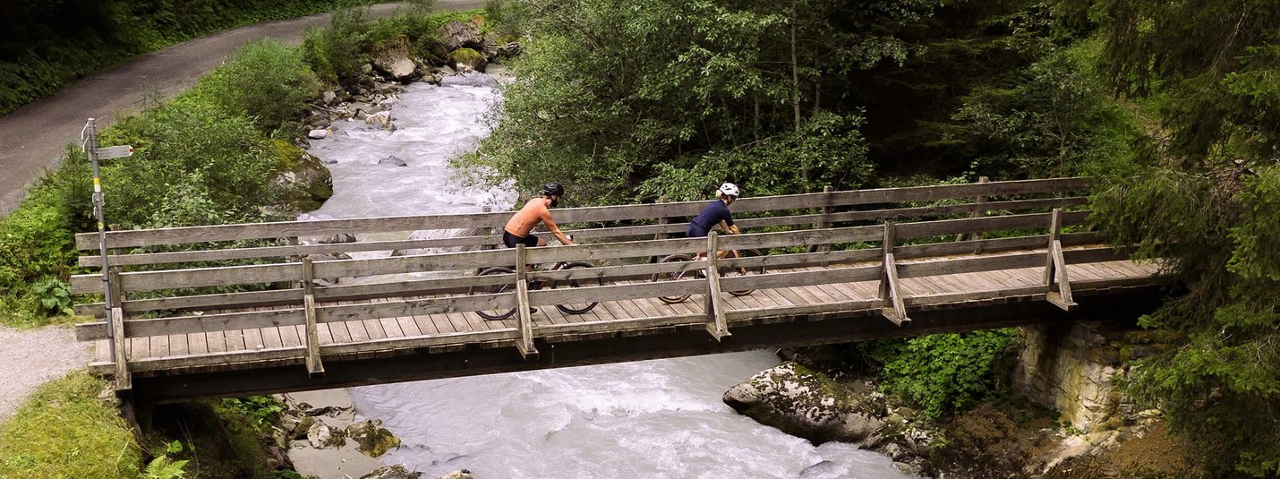 Graveltour Silbertal © Janine Brugger - Vorarlberg Tourismus Zwei Radfahrer fahren über eine Holzbrücke über einen Fluss in einem Wald