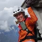 Smiling woman wearing helmet and orange jacket climbing a wooden structure in the mountains