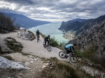 Northern Lake Garda © Gijs Ferkranus - Wielrenblad Magazine Three cyclists on a mountain trail overlooking a lake and mountains