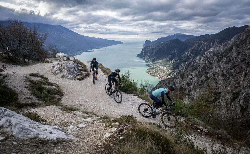 Gravel bike holiday on Northern Lake Garda © Gijs Ferkranus - Wielrenblad Magazine Three cyclists on a mountain trail overlooking a lake and mountains