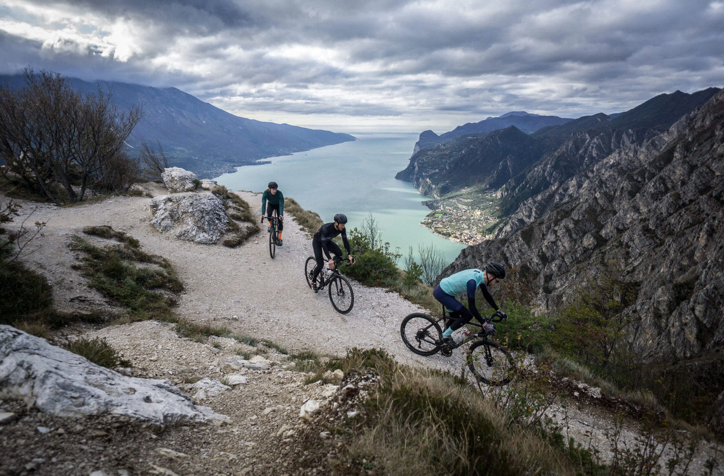 Italy © Gijs Ferkranus - Wielrenblad Magazine Three cyclists on a mountain trail overlooking a lake and mountains