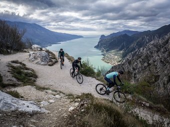 Drei Radfahrer auf einem Bergweg mit Blick auf einen See und Berge