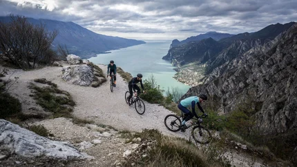 Three cyclists on a mountain trail overlooking a lake and mountains