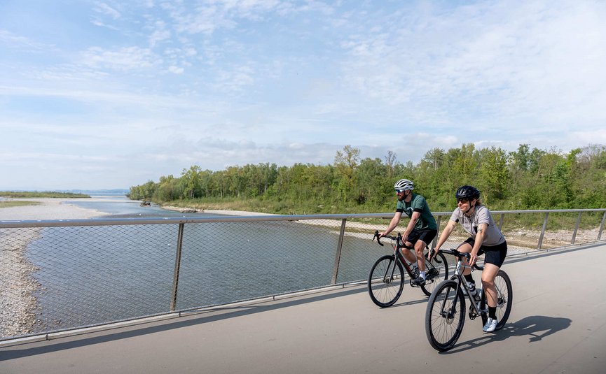 Zwei Radfahrer auf einer Brücke neben einem Fluss unter bewölktem Himmel