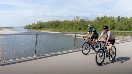 Two cyclists riding on a bridge beside a river under a partly cloudy sky