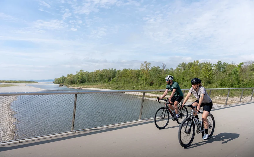 Two cyclists riding on a bridge beside a river under a partly cloudy sky