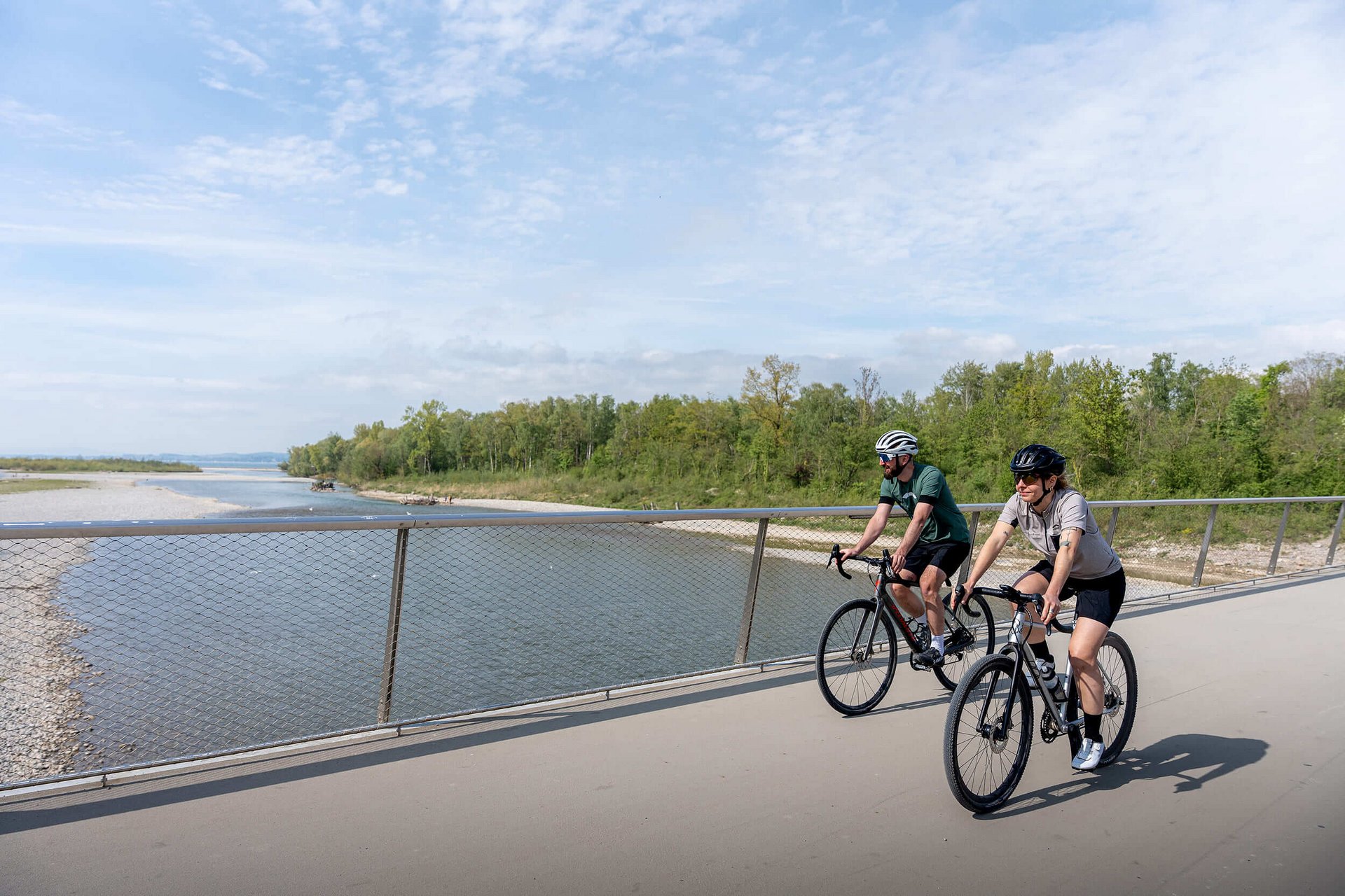 Gravelbike Holidays ยฉ Chris Gollhofer - Vorarlberg Tourismus Two cyclists riding on a bridge beside a river under a partly cloudy sky