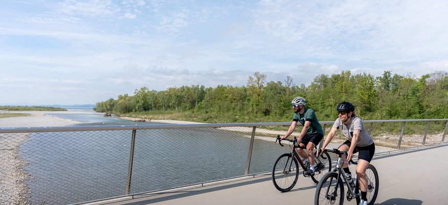 Zwei Radfahrer auf einer Brücke neben einem Fluss unter bewölktem Himmel