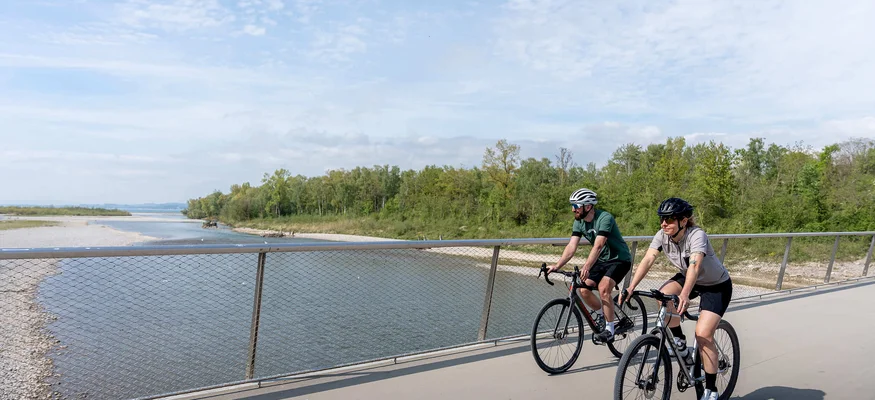Grenzenlos graveln in Vorarlberg © Chris Gollhofer - Vorarlberg Tourismus Zwei Radfahrer auf einer Brücke neben einem Fluss unter bewölktem Himmel