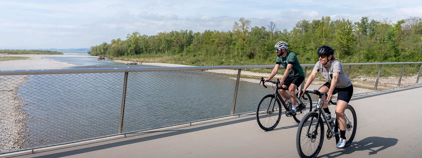 Graveltour Bregenz © Chris Gollhofer - Vorarlberg Tourismus Zwei Radfahrer auf einer Brücke neben einem Fluss unter bewölktem Himmel
