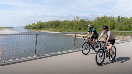 Graveltour Bregenz © Chris Gollhofer - Vorarlberg Tourismus Zwei Radfahrer auf einer Brücke neben einem Fluss unter bewölktem Himmel