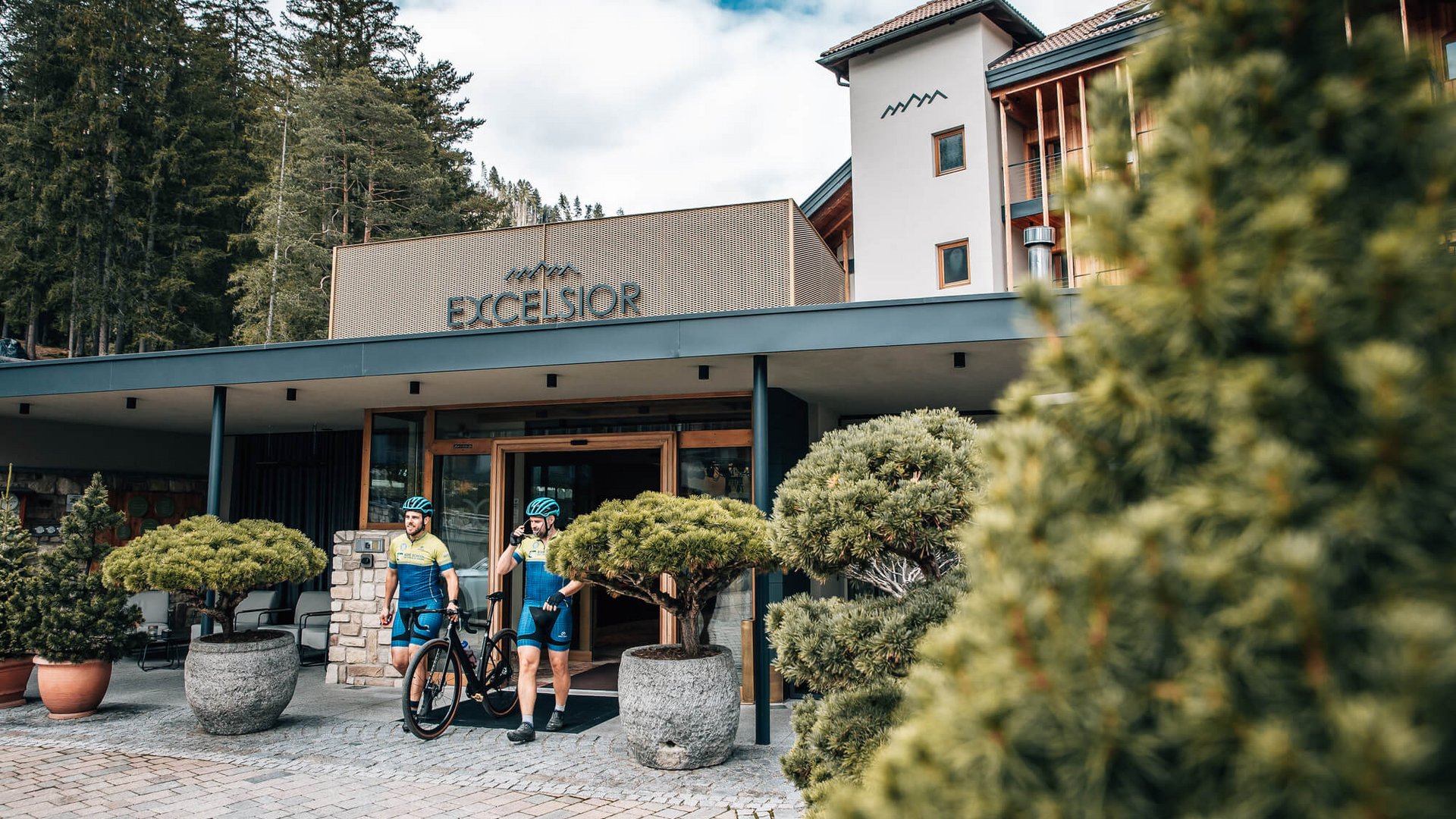 Zwei Radfahrer verlassen Hotel Excelsior in bewaldeter Berglandschaft