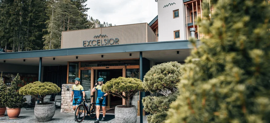 Herbst auf Schotter im Excelsior Dolomites Life Resort****s © Jens Scheibe Zwei Radfahrer verlassen Hotel Excelsior in bewaldeter Berglandschaft