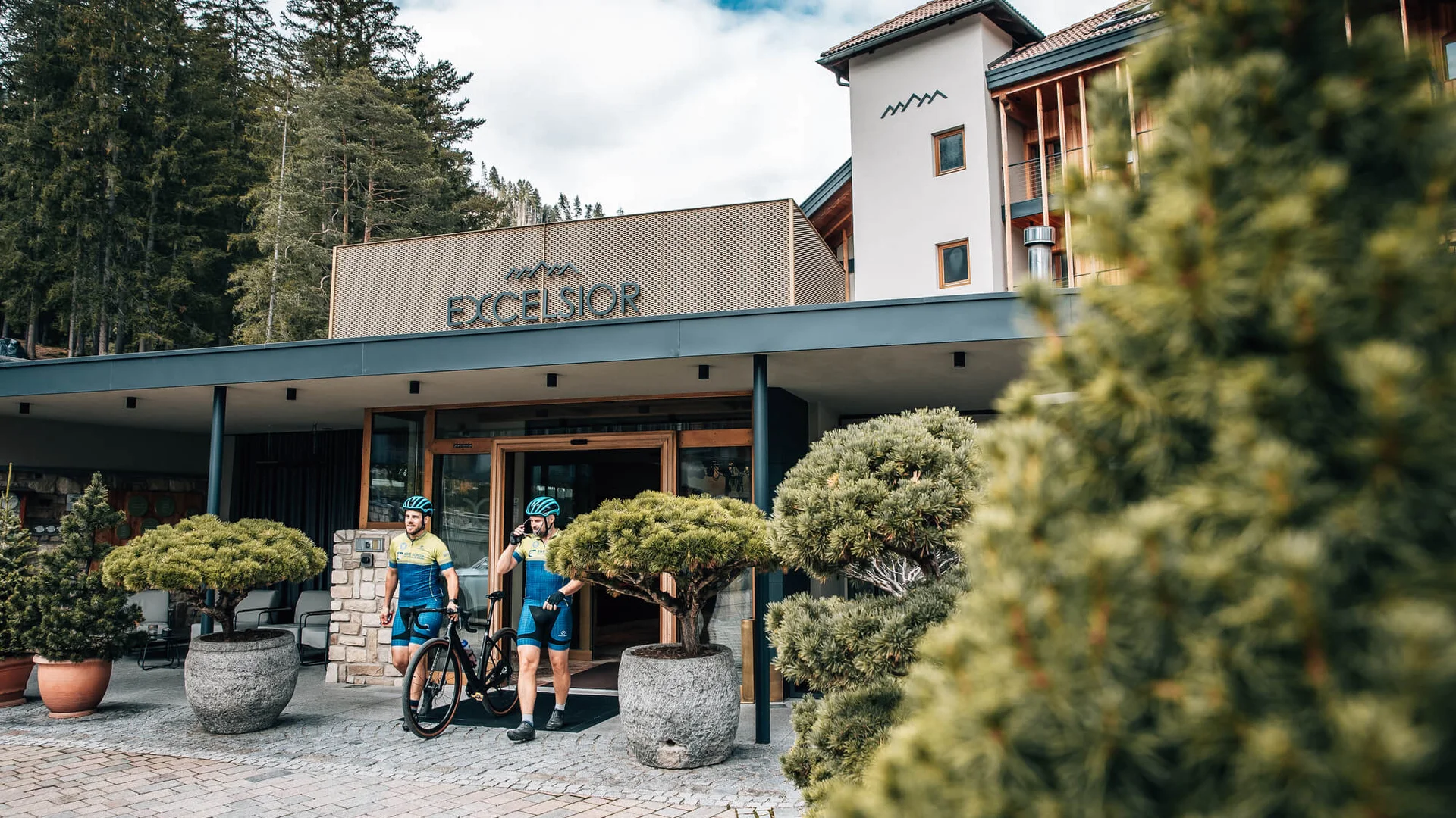 Zwei Radfahrer verlassen Hotel Excelsior in bewaldeter Berglandschaft