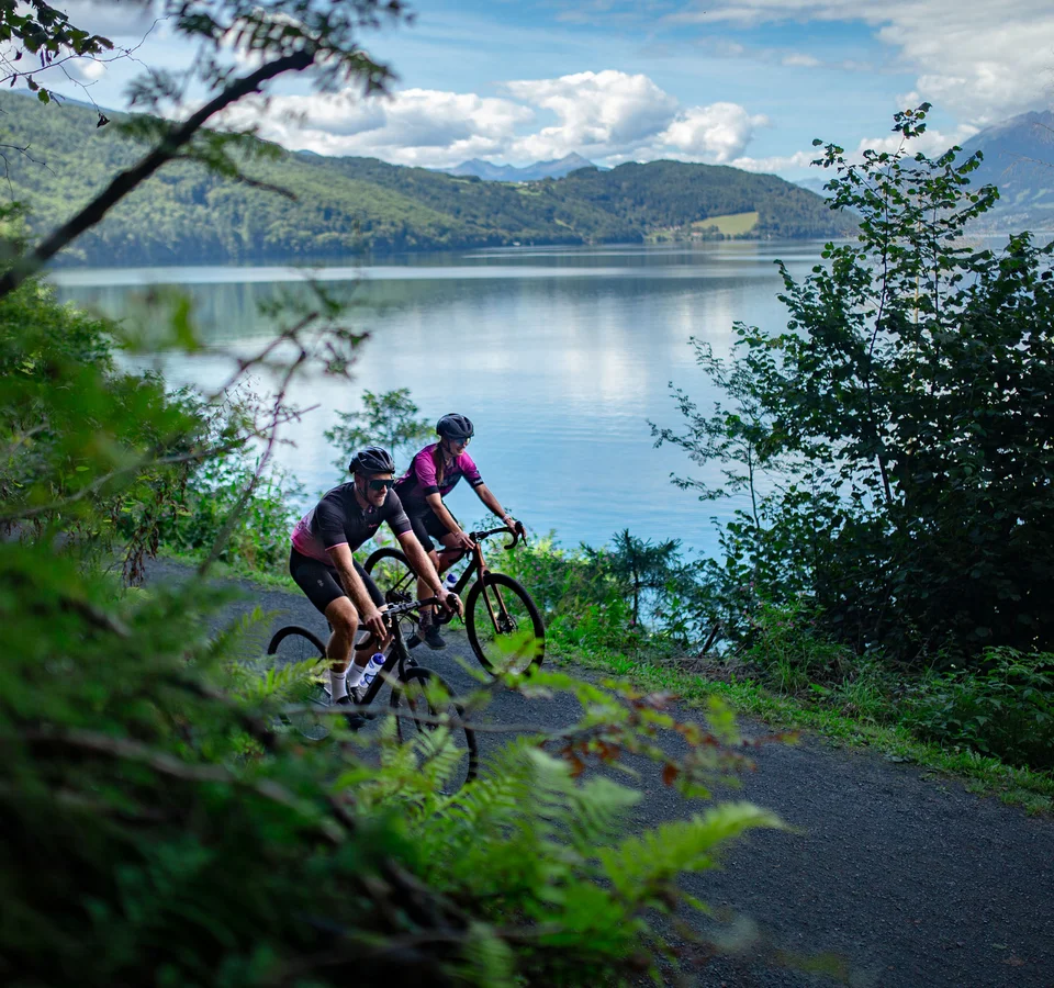 Zwei Radfahrer fahren auf einem Weg am See entlang mit Bergen im Hintergrund