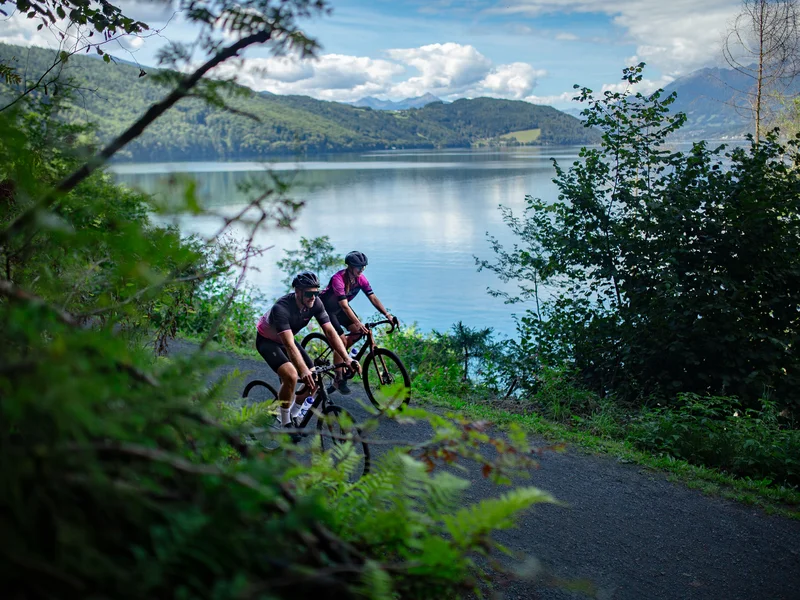 Zwei Radfahrer fahren auf einem Weg am See entlang mit Bergen im Hintergrund