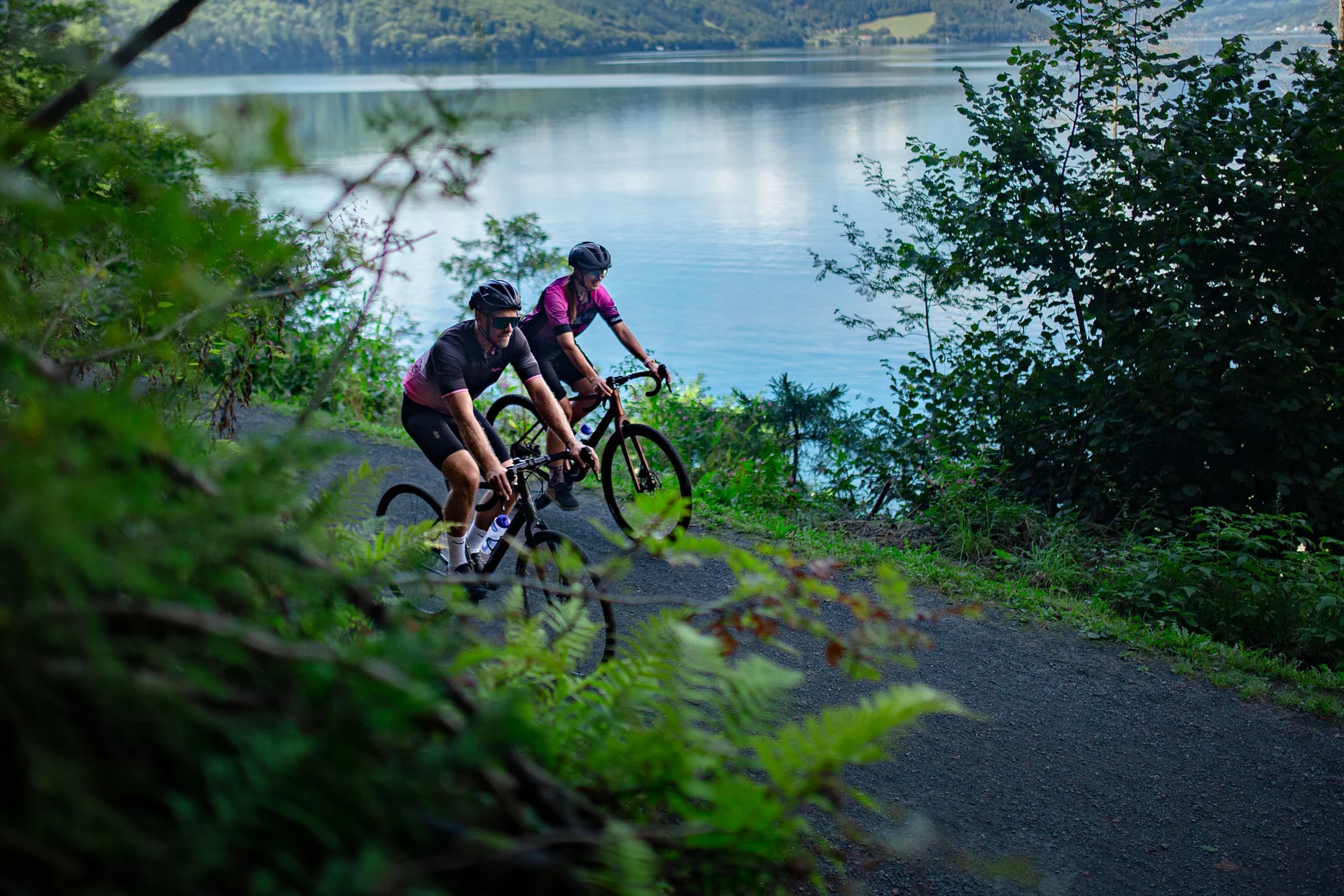 Gravelbike Holidays © Nicola Littmann Zwei Radfahrer fahren auf einem Weg am See entlang mit Bergen im Hintergrund