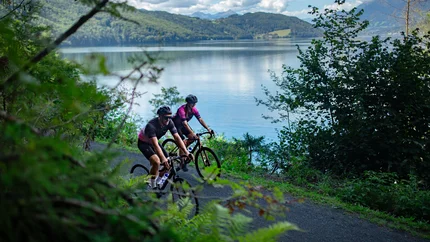 5 Gravelbike-Highlights in der NOCKBIKE-Area © Nicola Littmann Zwei Radfahrer fahren auf einem Weg am See entlang mit Bergen im Hintergrund