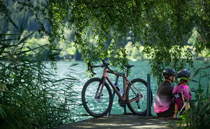 Two cyclists sitting by the lakeside with a bicycle under hanging trees