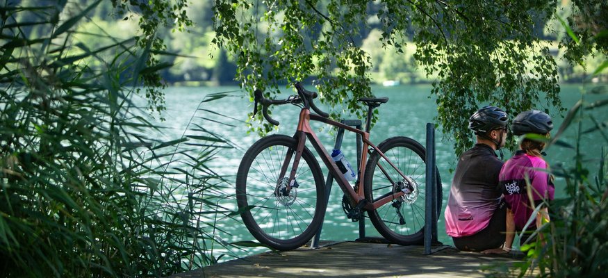 Two cyclists sitting by the lakeside with a bicycle under hanging trees