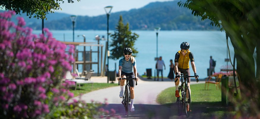 Zwei Radfahrer fahren auf einem Weg mit See und Blumen im Hintergrund