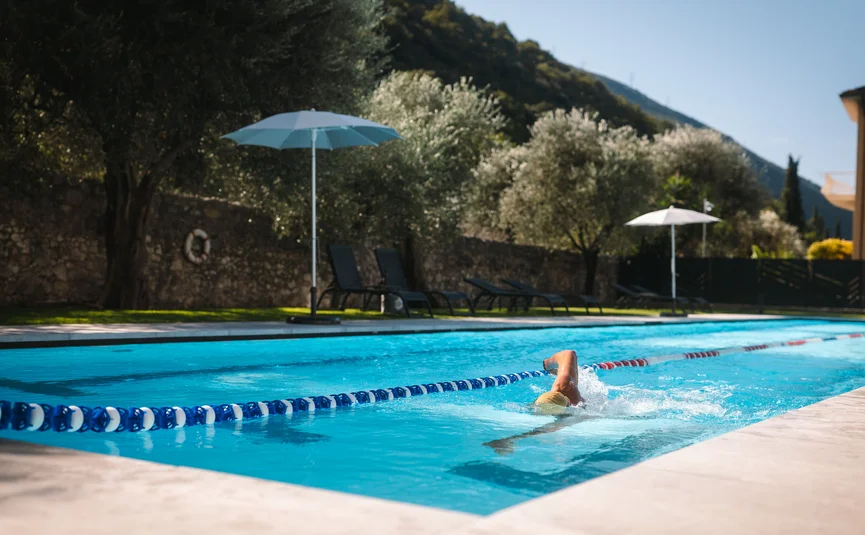 Person swimming in outdoor pool with sun umbrellas and trees in background