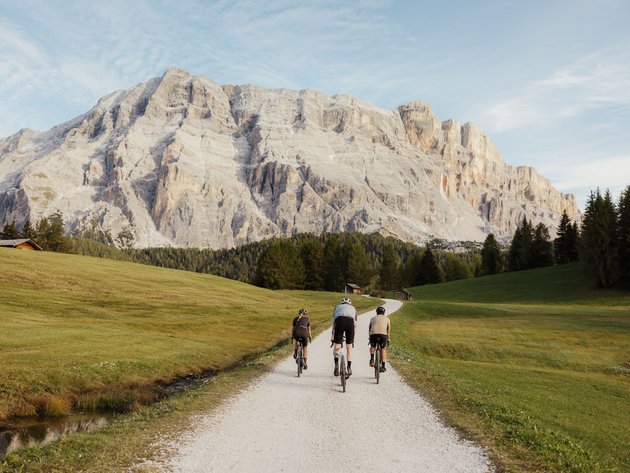 Alpine Gravel Experience © Alex Moling Drei Radfahrer auf einem Weg mit Berglandschaft und Wiesen