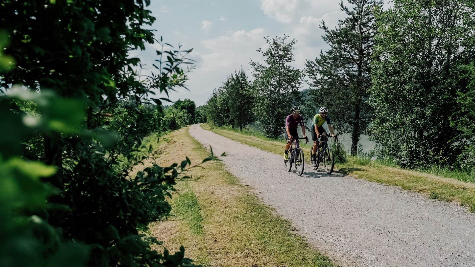 Zwei Radfahrer auf einem Waldweg neben einem See unter bewölktem Himmel