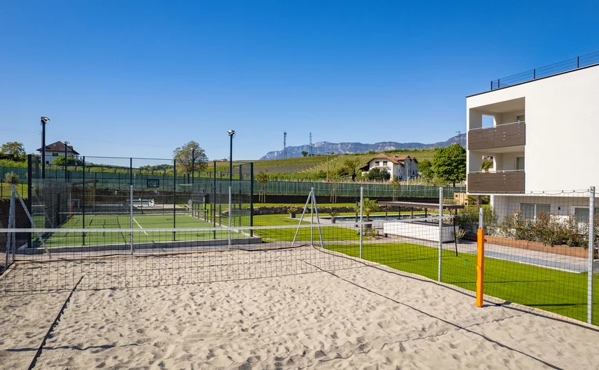 Sandy volleyball court next to tennis courts and modern building under clear sky
