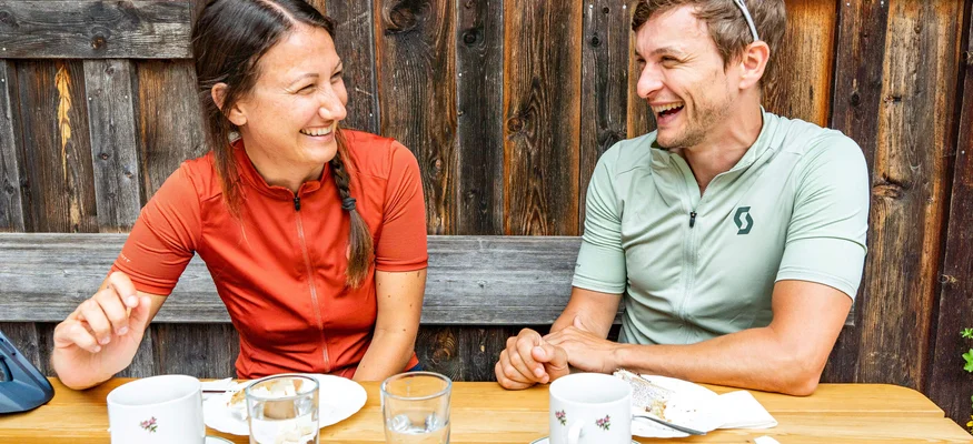 Zwei lachende Radfahrer sitzen draußen am Holztisch mit Kaffeetassen
