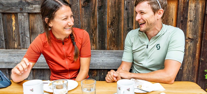 Zwei lachende Radfahrer sitzen draußen am Holztisch mit Kaffeetassen