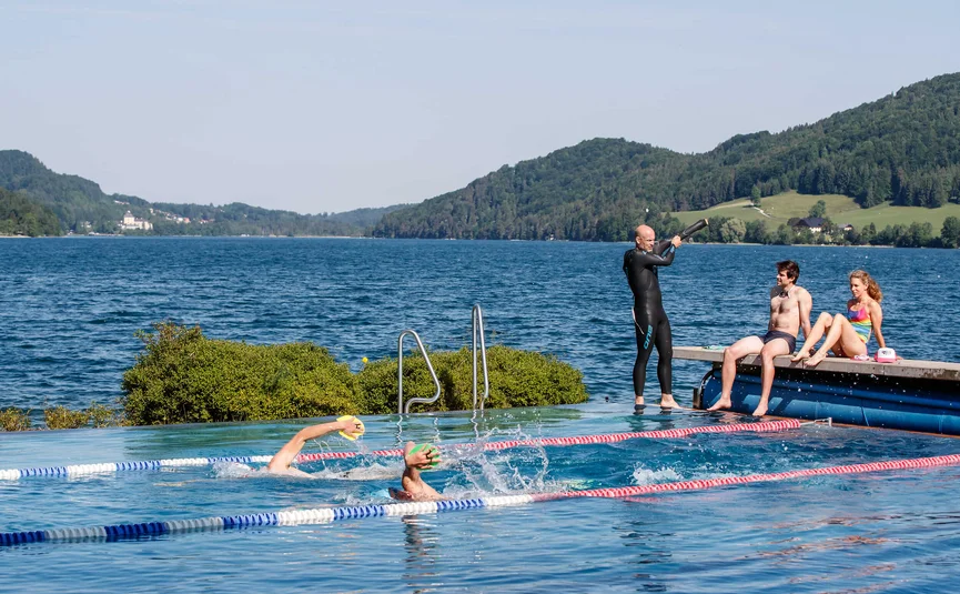People swimming in outdoor pool with mountains and lake in background