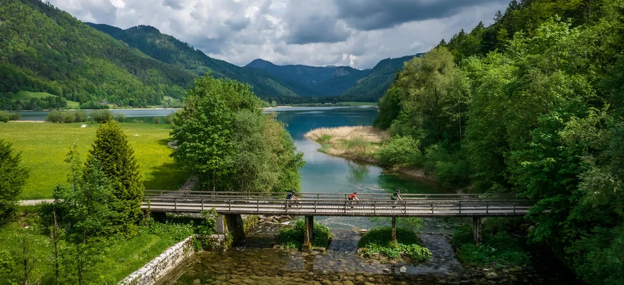 Cyclists on wooden bridge over river with green hills and cloudy sky